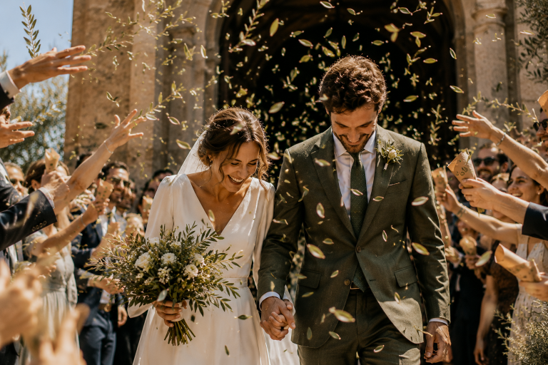 Novios saliendo de la ceremonia mientras familiares y amigos lanzan hojas de olivo frescas al aire en una celebración de boda al aire libre.